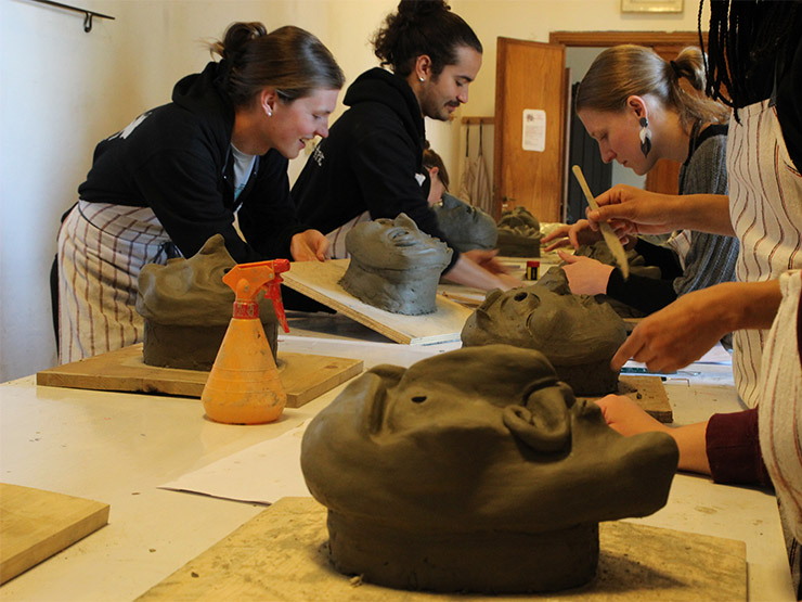 Several college-age students work at a table, carving traditional commedia dell'arte masks out of clay.